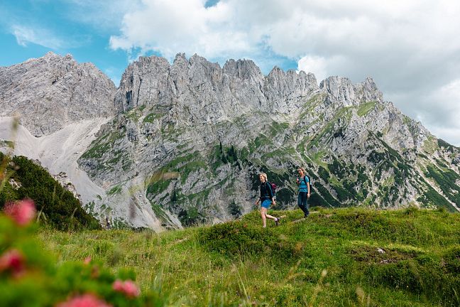 Zwei Wanderer spazieren auf einer grünen Wiese vor einem imposanten Bergmassiv in den Alpen. Der Himmel ist bewölkt, mit blauen Flecken sichtbar.