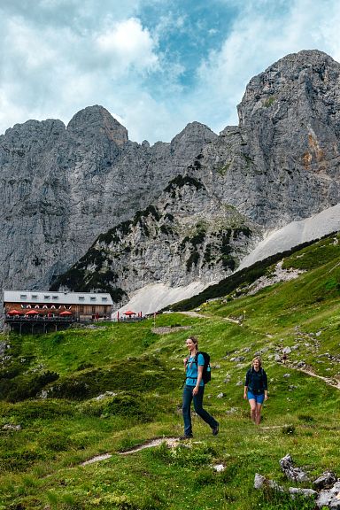 Ein Paar sitzt auf einer Bank vor einer beeindruckenden Bergkulisse am Wilden Kaiser, mit Wanderkleidung und Rucksäcken, umgeben von sommerlichem Grün.