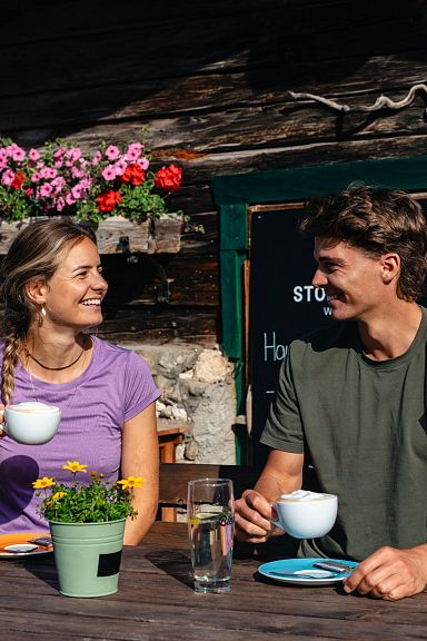 Ein Mann und eine Frau sitzen bei Sonnenschein lächelnd vor einer Berghütte und trinken Kaffee. Blumen schmücken die Hütte im Hintergrund.