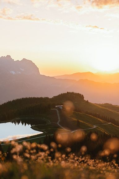 Sonnenuntergang über einer bergigen Landschaft mit einem kleinen See im Vordergrund und Silhouetten von Bergspitzen im Hintergrund.