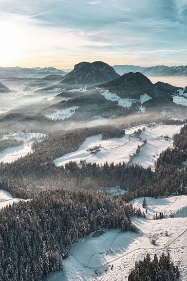 A snowy landscape with mountains and valleys under a bright blue sky, featuring the Wilder Kaiser range in the middle distance.