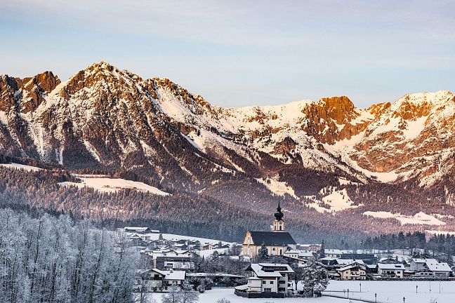 Verschneite Berglandschaft mit einer Kunstinstallation aus hohen, schmalen Steinsäulen und einer stilisierten Sonne im Hintergrund vor blauem Himmel.