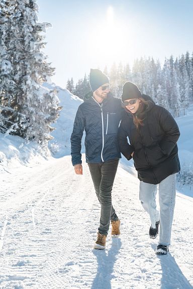 Zwei Menschen spazieren lachend auf einem verschneiten Weg in einer winterlichen Landschaft mit Sonnenschein, umgeben von schneebedeckten Bäumen.