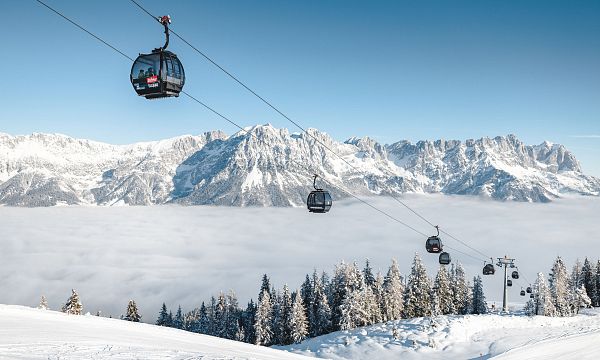 Schneebedeckte Berglandschaft mit Seilbahn und Gondeln über einer Wolkendecke, umgeben von verschneiten Bäumen und majestätischen Alpen im Hintergrund.