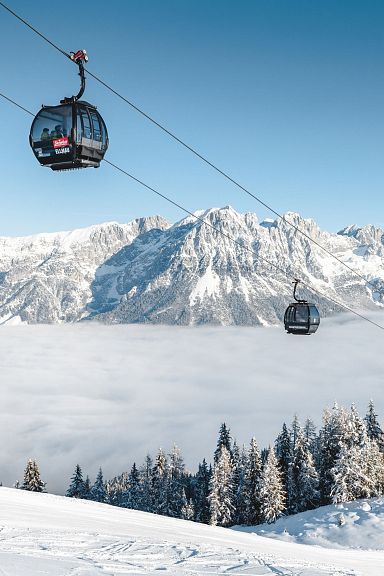 Schneebedeckte Berglandschaft mit Seilbahn und Gondeln über einer Wolkendecke, umgeben von verschneiten Bäumen und majestätischen Alpen im Hintergrund.