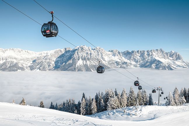 Schneebedeckte Berglandschaft mit Seilbahn und Gondeln über einer Wolkendecke, umgeben von verschneiten Bäumen und majestätischen Alpen im Hintergrund.