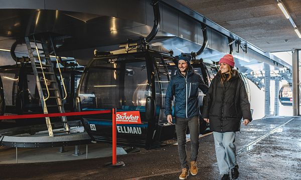 Zwei Personen gehen fröhlich neben einer Seilbahnstation in Ellmau, Tirol. Im Hintergrund sind moderne Gondeln der SkiWelt Wilder Kaiser zu sehen.