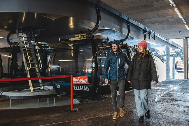 Couple walks inside Ellmau cable car station, part of Austria's Wilder Kaiser region, with SkiWelt sign visible, indicating a popular skiing destination.