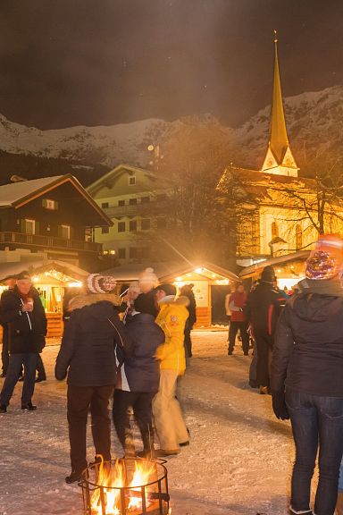 Menschen genießen einen winterlichen Weihnachtsmarktabend in einem verschneiten Dorf mit beleuchteten Holzbuden und Bergkulisse im Hintergrund.