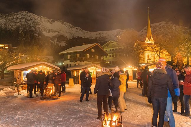 A winter market at night with the illuminated stalls, a church, and snow-covered mountains in the background of the Wilder Kaiser area.