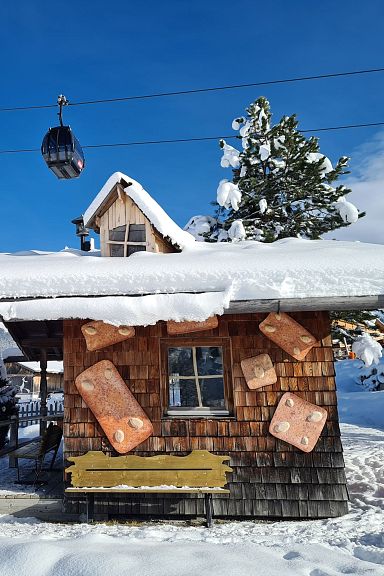 Verschneite Holzhütte mit rustikalem Charme, umgeben von Winterlandschaft. Eine Seilbahn schwebt am blauen Himmel über der Hütte. Idylle in den Alpen.