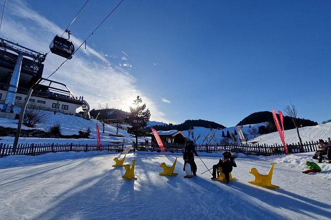 Kinder spielen auf einem Holzspielgerät im Schnee bei sonnigem Winterwetter. Im Hintergrund sind schneebedeckte Hügel und ein Gebäude zu sehen.