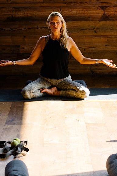Eine Frau sitzt meditierend in einem Yoga-Studio auf einer Matte, umgeben von Teilnehmern, die sich auf dem Boden entspannen. Warme Holzwände schaffen ein gemütliches Ambiente.