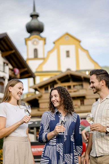 Drei Personen stehen lächelnd vor traditionellen Tiroler Gebäuden mit malerischem Kirchturm im Hintergrund und halten Weingläser in der Hand.