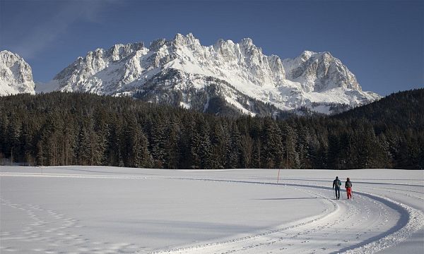 Verschneite Landschaft mit einem majestätischen Bergmassiv im Hintergrund. Zwei Personen laufen auf einem schneebedeckten Weg durch eine winterliche Waldszenerie.