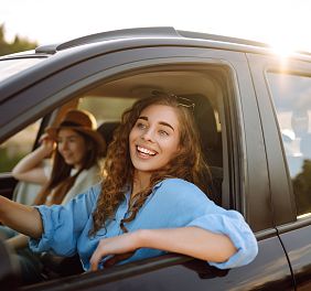 Zwei junge Frauen sitzen in einem Auto. Die Fahrerin lächelt entspannt, während die Sonne durch das Fenster scheint, und eine Passagierin sitzt im Hintergrund.