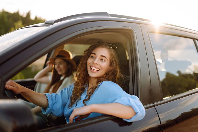 Two people sitting in a parked car, enjoying the sunlight. The person in front is smiling and leaning out of the window, while the other sits in the passenger seat.