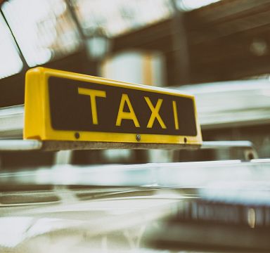 Nahaufnahme eines Taxis mit gelbem "Taxi"-Schild auf dem Dach, unscharfer Hintergrund von weiteren Fahrzeugen sichtbar, städtische Umgebung.