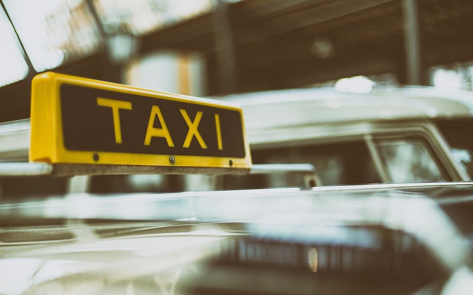 A vintage taxi with a yellow "TAXI" sign on top, parked in a sunlit area with blurred surroundings, creating a nostalgic atmosphere.