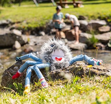 Buntes Stofftier in Spinnenform mit lächelndem Gesicht auf einem Felsen; im Hintergrund spielen Kinder an einem Bach bei sonnigem Wetter.