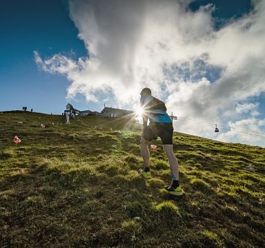 Ein Läufer erklimmt einen grünen Hügel bei Sonnenschein, während im Hintergrund Seilbahnen zu sehen sind. Der Himmel ist blau mit einigen Wolken.