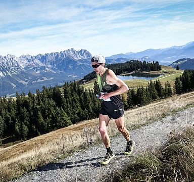Ein Läufer in Sportkleidung joggt auf einem Pfad mit einer Berglandschaft im Hintergrund. Der Himmel ist klar, und die umliegenden Wälder sind grün.