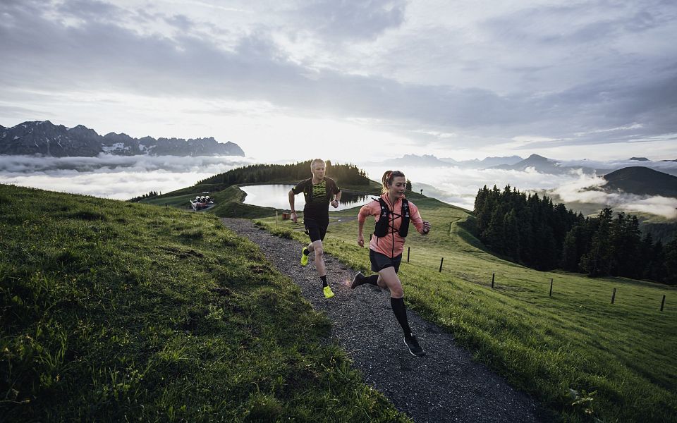 Zwei Läufer auf einem Bergweg, umgeben von grünen Wiesen und Bergen im Hintergrund. Der Himmel ist bewölkt und es liegt Nebel in den Tälern.