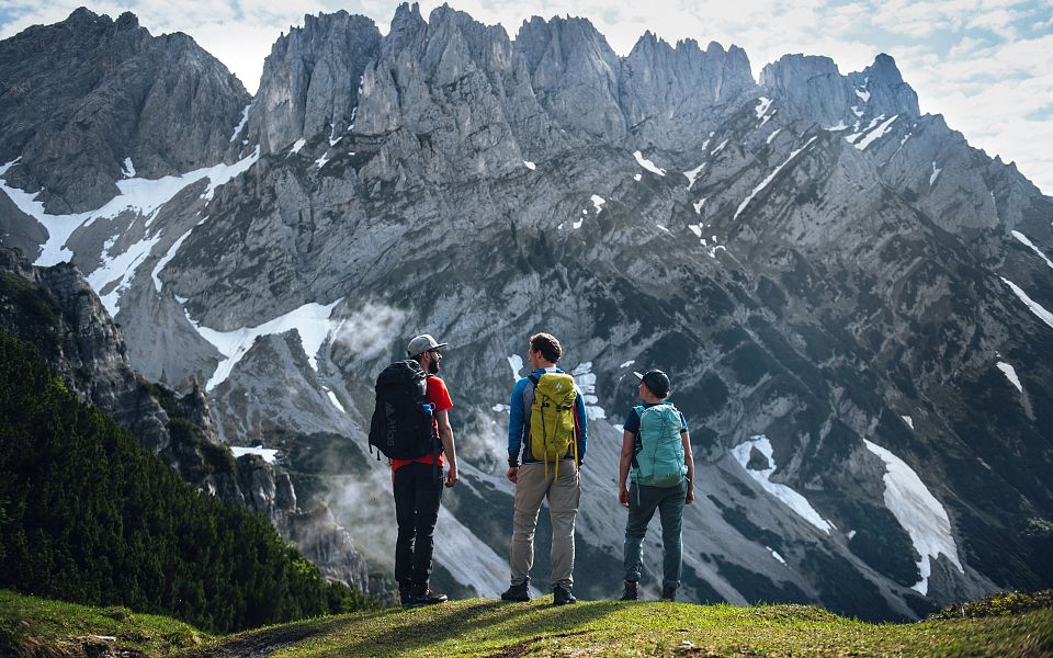 Drei Wanderer stehen vor einer beeindruckenden Bergkulisse in den Alpen, umgeben von schneebedeckten Gipfeln und dramatischen Felsformationen.