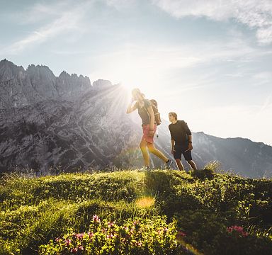 Zwei Wanderer auf einem blühenden Bergpfad, Sonnenstrahlen brechen durch die Wolken, im Hintergrund ragt ein imposantes Bergmassiv in den Himmel.