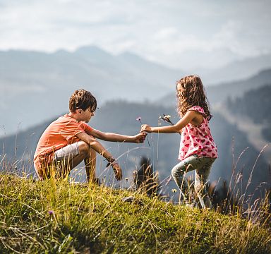 Zwei Kinder spielen auf einer Wiese in den Bergen. Der Junge kniet, während das Mädchen Blumen hält. Im Hintergrund sind nebelige Berge zu sehen.