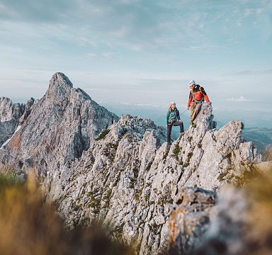 Zwei Bergsteiger klettern auf einem felsigen Grat im Gebirge unter blauem Himmel. Im Hintergrund sind steile Felsformationen und Täler zu sehen.