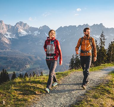 Zwei Menschen wandern auf einem Pfad mit Blick auf das beeindruckende Bergmassiv des Wilden Kaisers in Tirol. Die Sonne scheint, der Himmel ist blau.