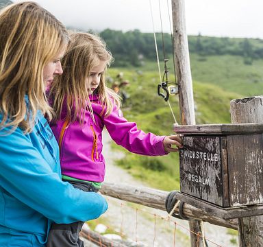 Eine Frau hält ein Kind, das neugierig eine hölzerne Schilderbox im Grünen betrachtet. Beide tragen bunte Outdoorjacken. Im Hintergrund sind Berge zu sehen.
