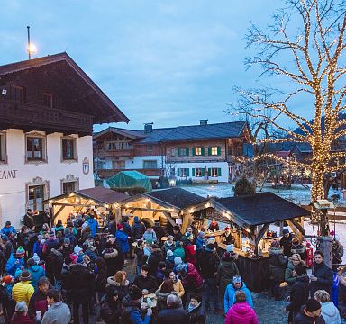 Crowded Christmas market scene with festive lights, wooden stalls, a tree wrapped in lights, and traditional Alpine buildings in a charming village setting.