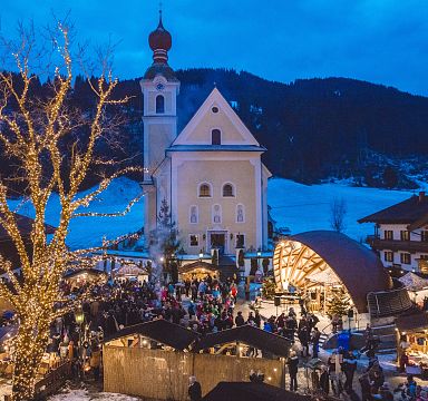 Ein festlich beleuchteter Weihnachtsmarkt vor einer Kirche in einem verschneiten Dorf bei Abenddämmerung, mit vielen Besuchern und dekorierten Ständen.
