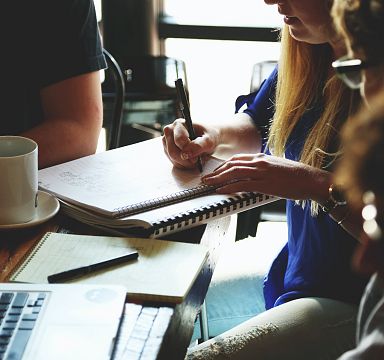 Group of people collaborating at a table with notebooks, a laptop, and a cup of coffee, engaging in a discussion or brainstorming session.