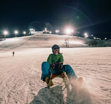 Person beim Nachtrodeln auf einer beleuchteten Skipiste, umgeben von Schneelandschaft. Im Hintergrund sind weitere Skisportler zu sehen.