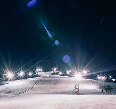 Nachts beleuchtete Skipiste mit klaren Himmel und kleinen Lichtern in der Ferne. Der Schnee reflektiert das Licht, was eine magische Winteratmosphäre schafft.