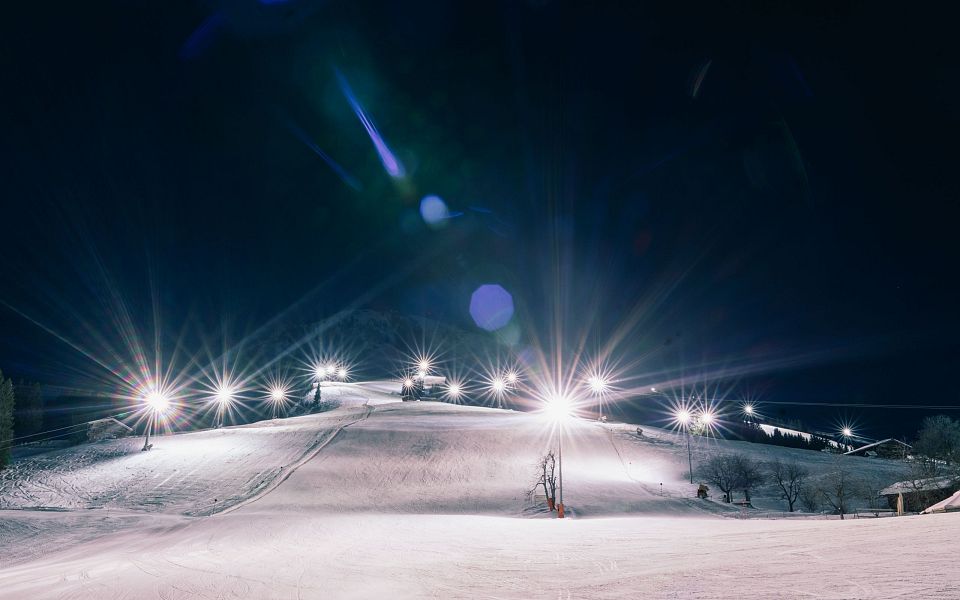 Nachts beleuchtete Skipiste mit klaren Himmel und kleinen Lichtern in der Ferne. Der Schnee reflektiert das Licht, was eine magische Winteratmosphäre schafft.