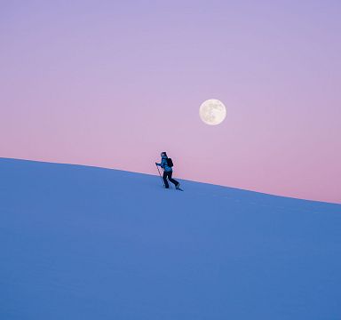 Eine Person wandert bei Sonnenuntergang in einer verschneiten Landschaft, der Vollmond steht am Himmel. Der Horizont zeigt zarte Blau- und Lilatöne.