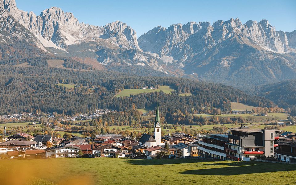 Panoramablick auf eine Tiroler Ortschaft vor einem imposanten Bergmassiv bei klarem Himmel, umgeben von grünen Wiesen und dichten Wäldern.