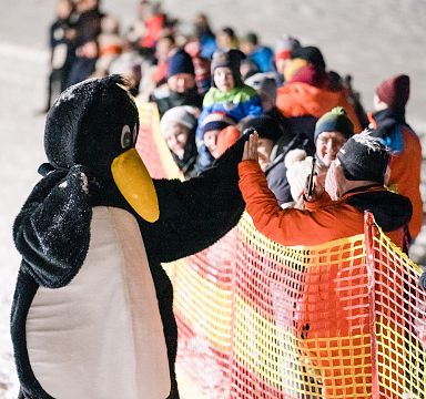 A penguin mascot interacts with people in winter clothing at a snowy event with bright lights in the Wilder Kaiser region.