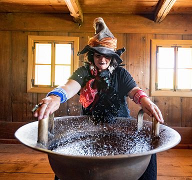 Frau in traditioneller Kleidung mit Hut steht in rustikaler Holzhütte und streut Körner in eine große Schale. Die Szene wirkt traditionell und authentisch.