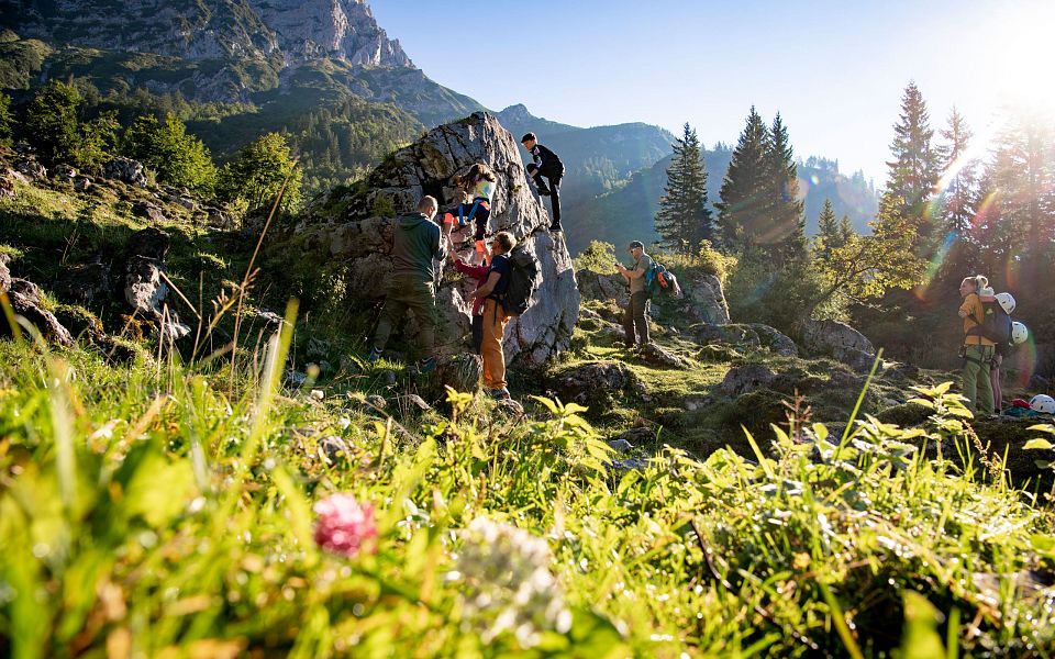 Ein sonniger Tag in den Alpen, Menschen klettern an einem Felsen inmitten grüner Wiesen und Bäume, im Hintergrund erhebt sich ein imposantes Bergmassiv.