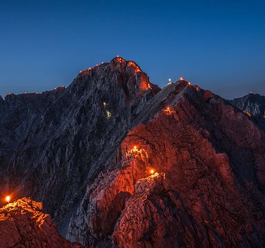 Bergkette bei Nacht mit beleuchteten Gipfelkreuzen und Wegen, die durch Fackeln oder Lichter markiert sind. Der Himmel ist dunkelblau und klar.