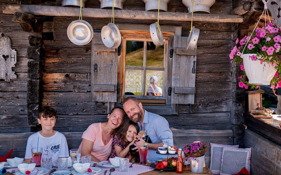Familie sitzt lächelnd an einem Holztisch vor einer Almhütte. Über ihnen hängen Milchkannen. Blumenschmuck und Wanddeko verleihen eine gemütliche Atmosphäre.