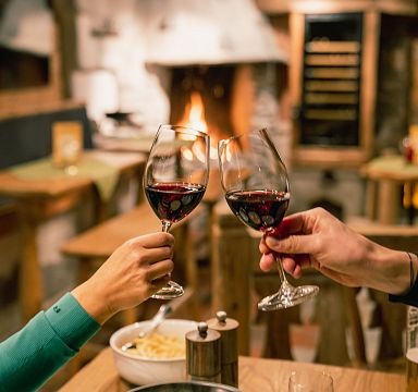 Zwei Personen stoßen mit Weingläsern an einem gemütlichen Holz-Tisch vor einem Kamin an, im Hintergrund ein warmes, einladendes Ambiente.