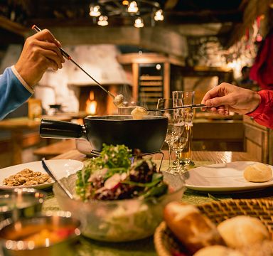Zwei Personen genießen Fondue in einer gemütlichen Alpenhütte. Auf dem Tisch stehen Brot, Salat und Dips. Eine warme, einladende Atmosphäre herrscht vor.
