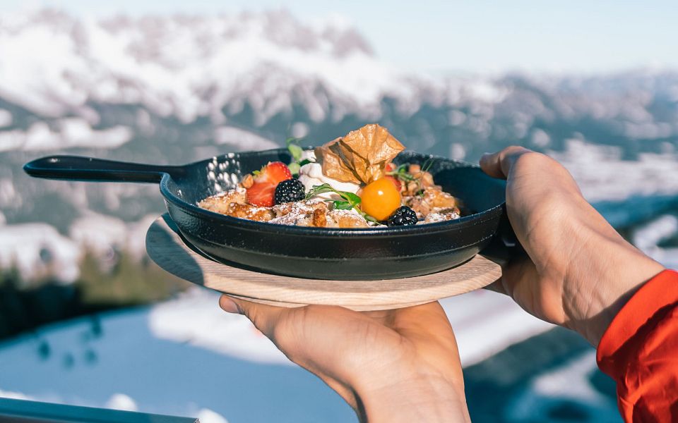 Eine Pfanne mit Kaiserschmarrn, serviert vor einer verschneiten Berglandschaft bei klarem Himmel. Der winterliche Hintergrund bietet eine beeindruckende Aussicht.