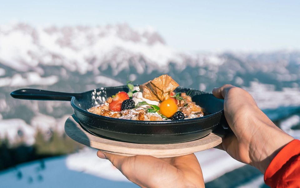 A delicious Tyrolean dish in a pan is held up with the snowy Wilder Kaiser mountains in the background, showcasing a blend of culinary and natural beauty.